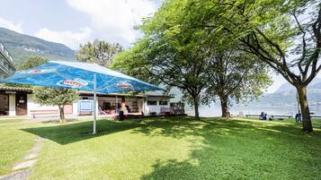 Une zone herbeuse avec un parasol bleu, un bâtiment et un arbre au bord d’un lac.