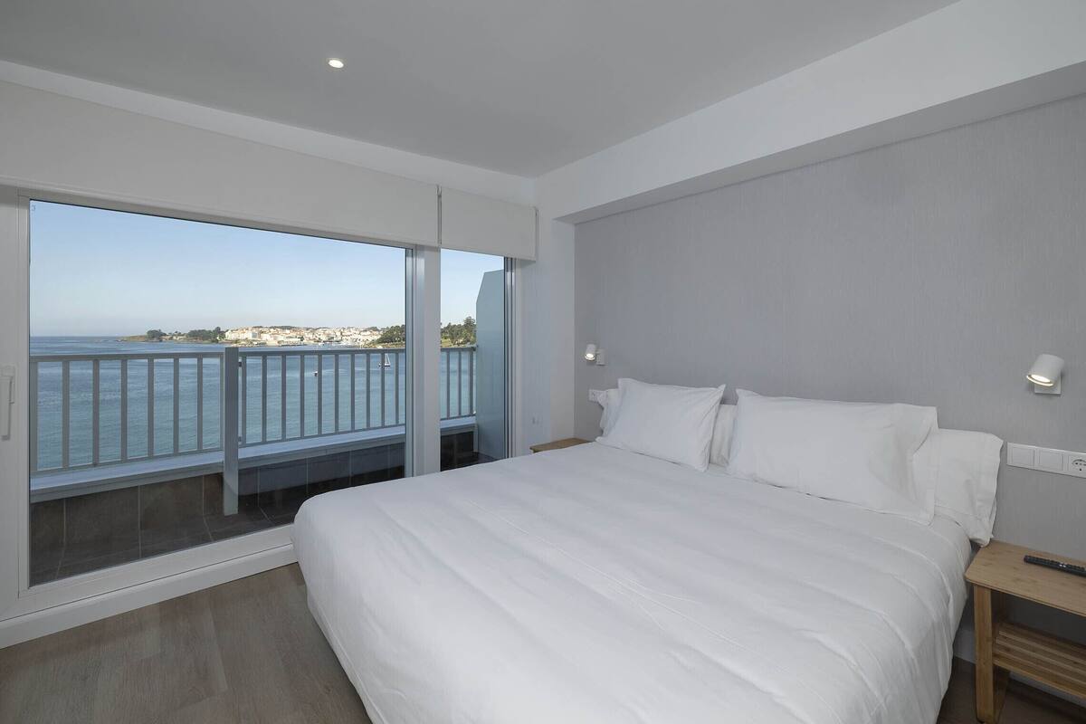 Quarto moderno com cama grande, mesa de cabeceira e vista para o mar através de uma porta de correr de vidro.