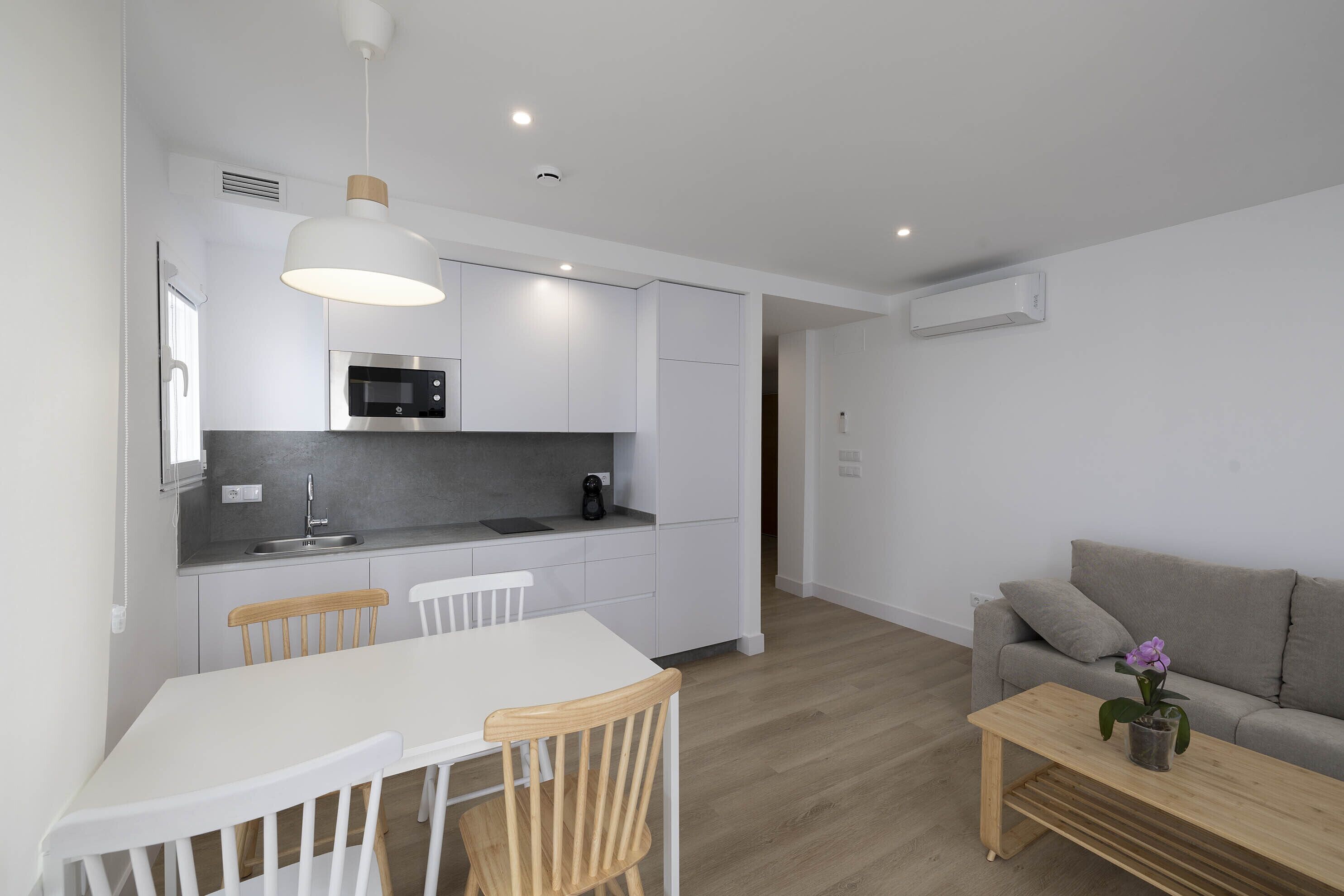 A modern kitchen with white cabinets, a microwave, and a sink. There is a dining table with chairs and a wooden bench. A gray sofa and a small table with a potted plant are in the living area.