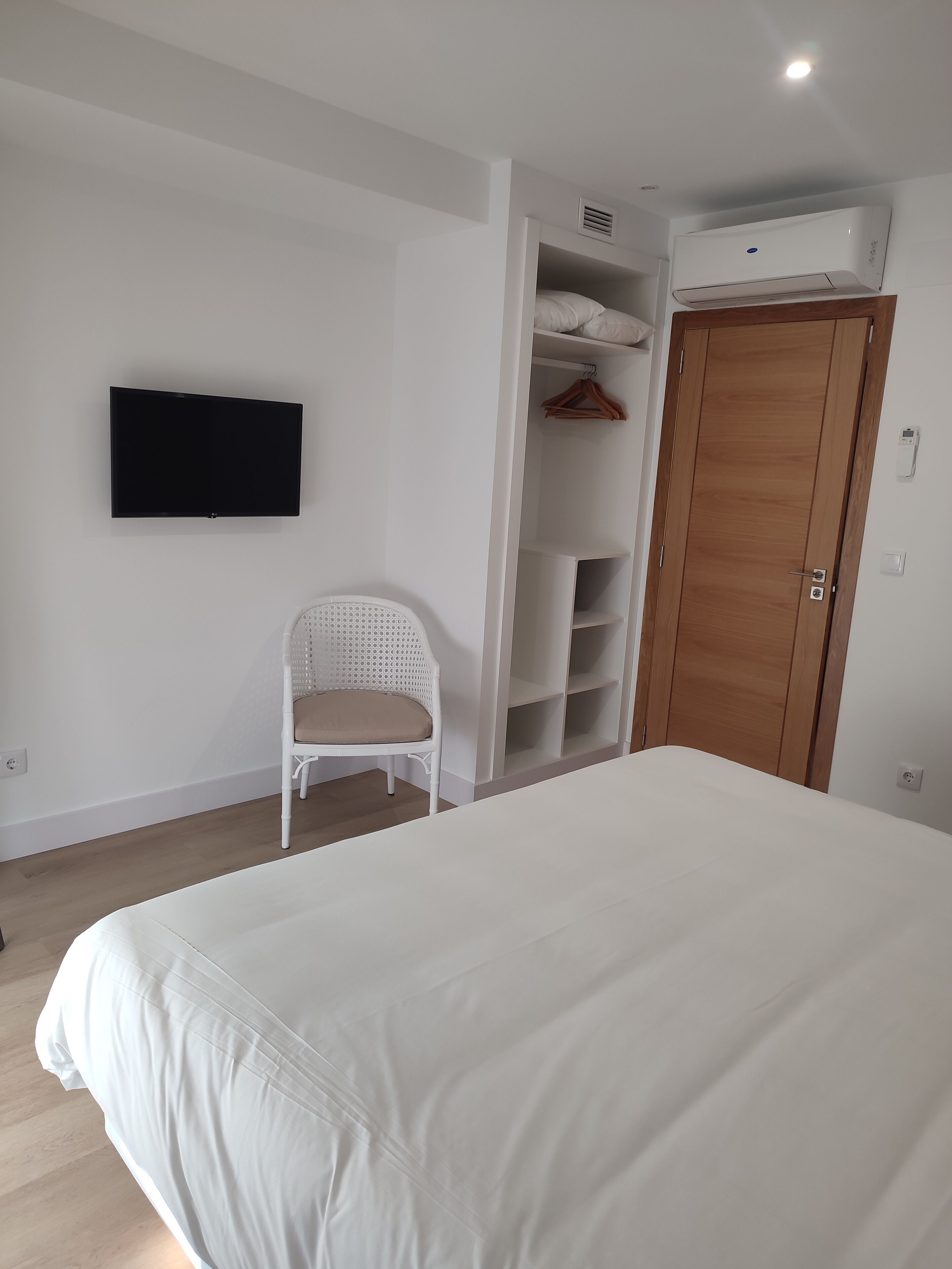Un dormitorio con una cama, una silla blanca, un armario y un televisor montado.