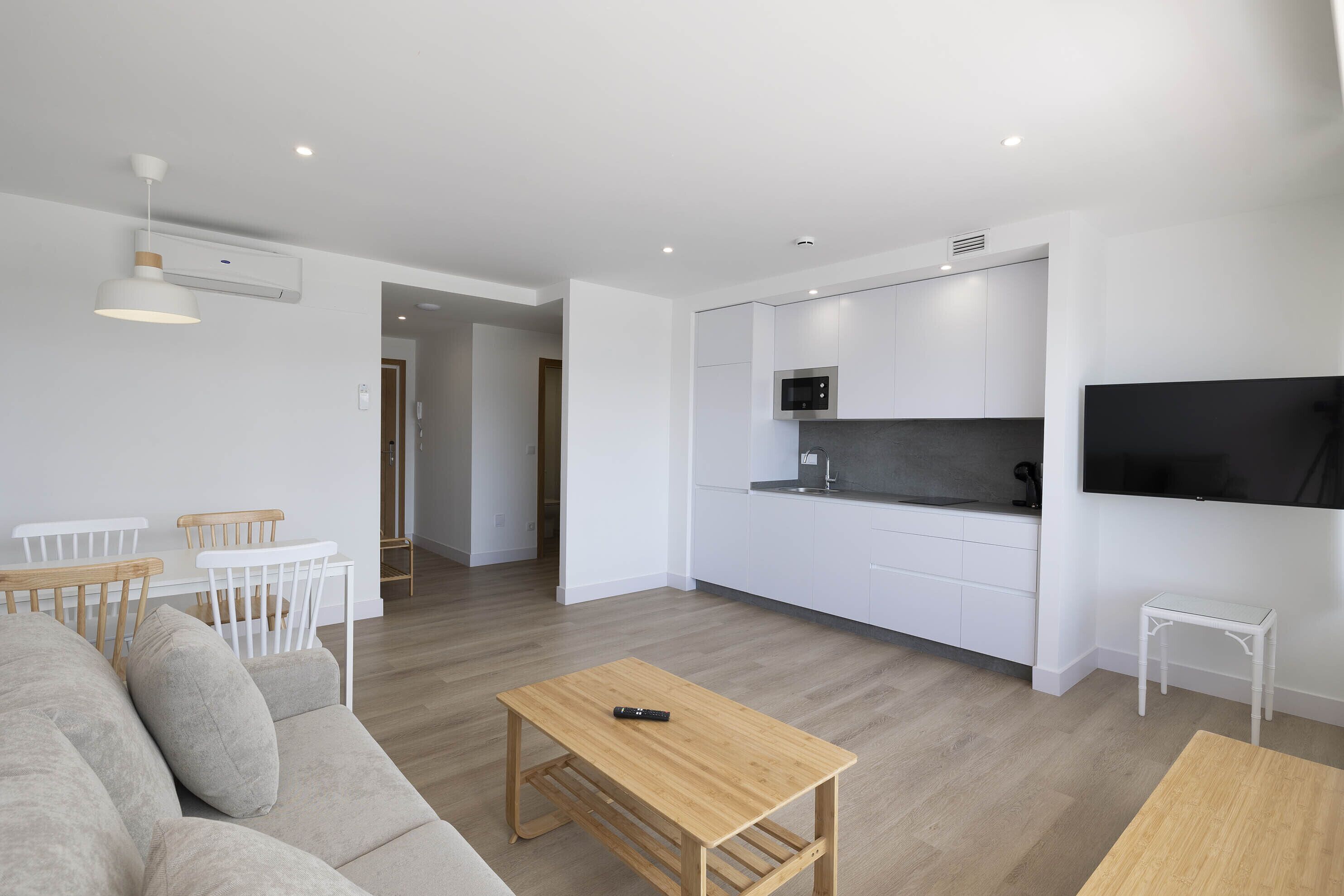 A modern living room with a sofa, a coffee table, and a dining area with chairs. There is a kitchen area with white cabinets and a microwave.