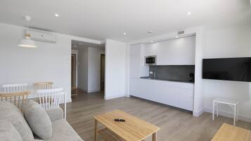 A modern living room with a sofa, a coffee table, and a dining area with chairs. There is a kitchen area with white cabinets and a microwave.