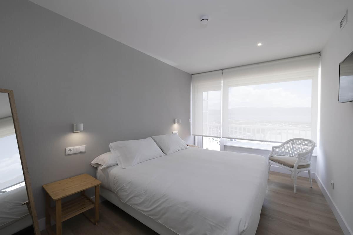 Quarto moderno com uma cama grande, uma mesinha de cabeceira de madeira, uma cadeira branca e vista para a cidade pela janela.