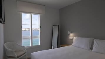 A hotel room with a bed, a chair, a window with a view of the sea, and a mirror on the wall.