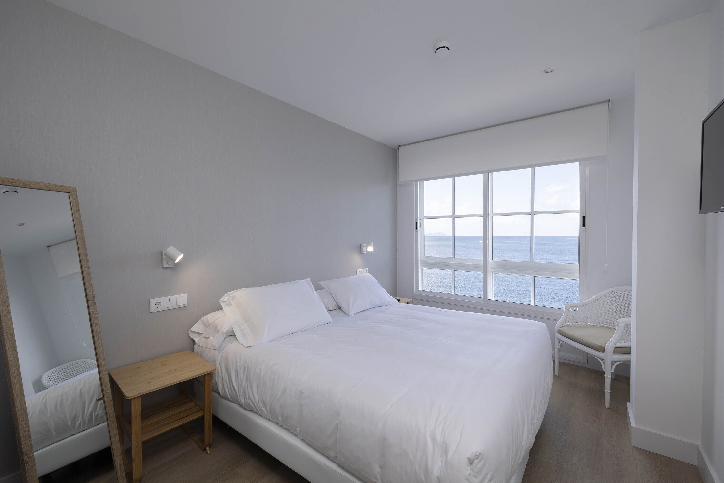 A bedroom with a large bed, a bedside table, a chair, a window with a view of the sea, and a mounted TV.