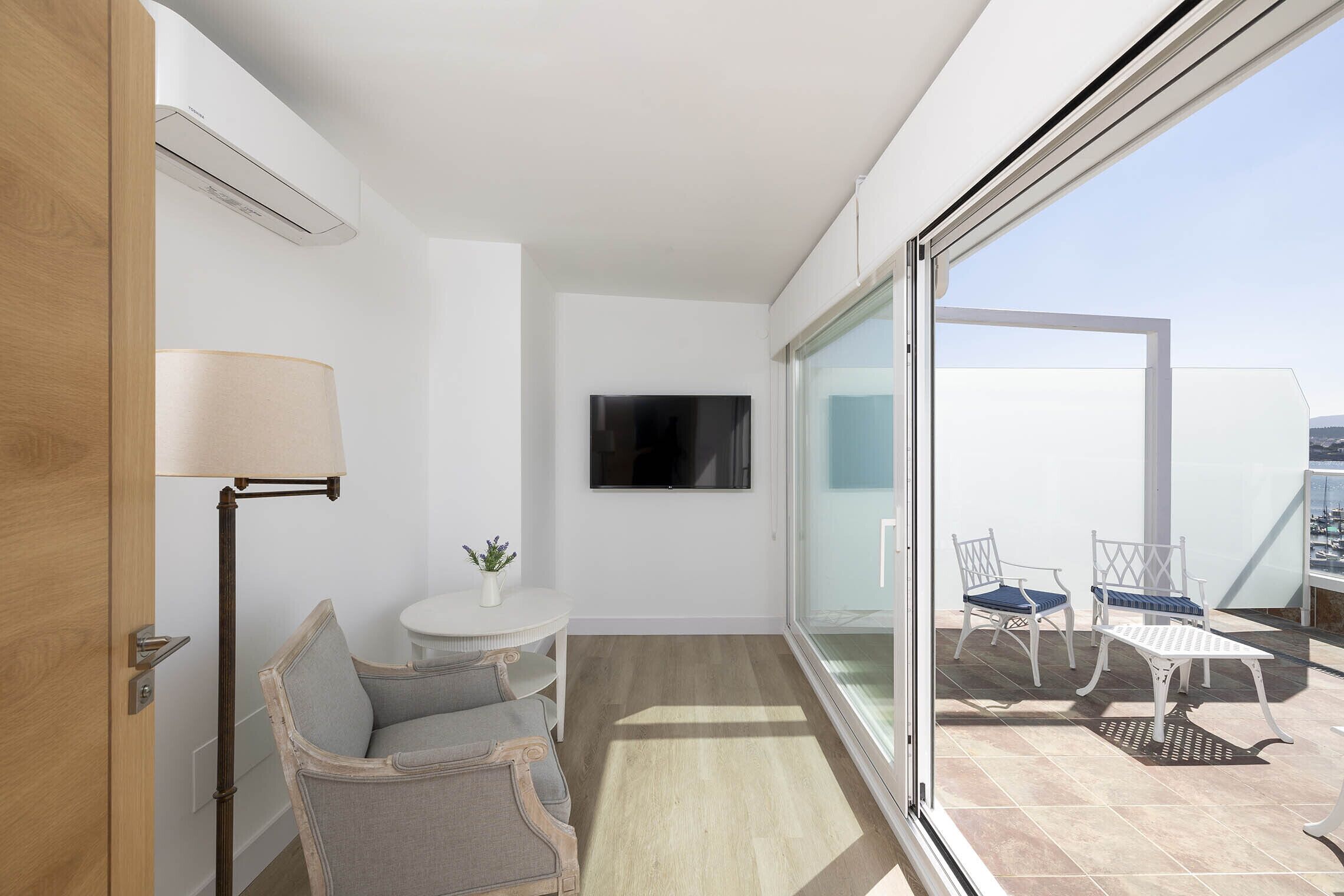 Una habitación moderna con puerta corrediza de vidrio que da a un balcón, un televisor de pantalla plana en la pared, una pequeña mesa redonda con una lámpara y dos sillas.
