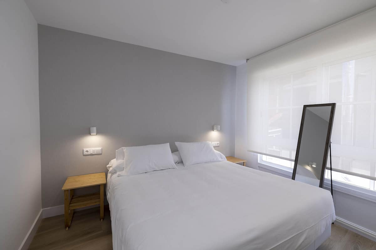 Quarto com cama, mesas de cabeceira, espelho e janela com persianas.