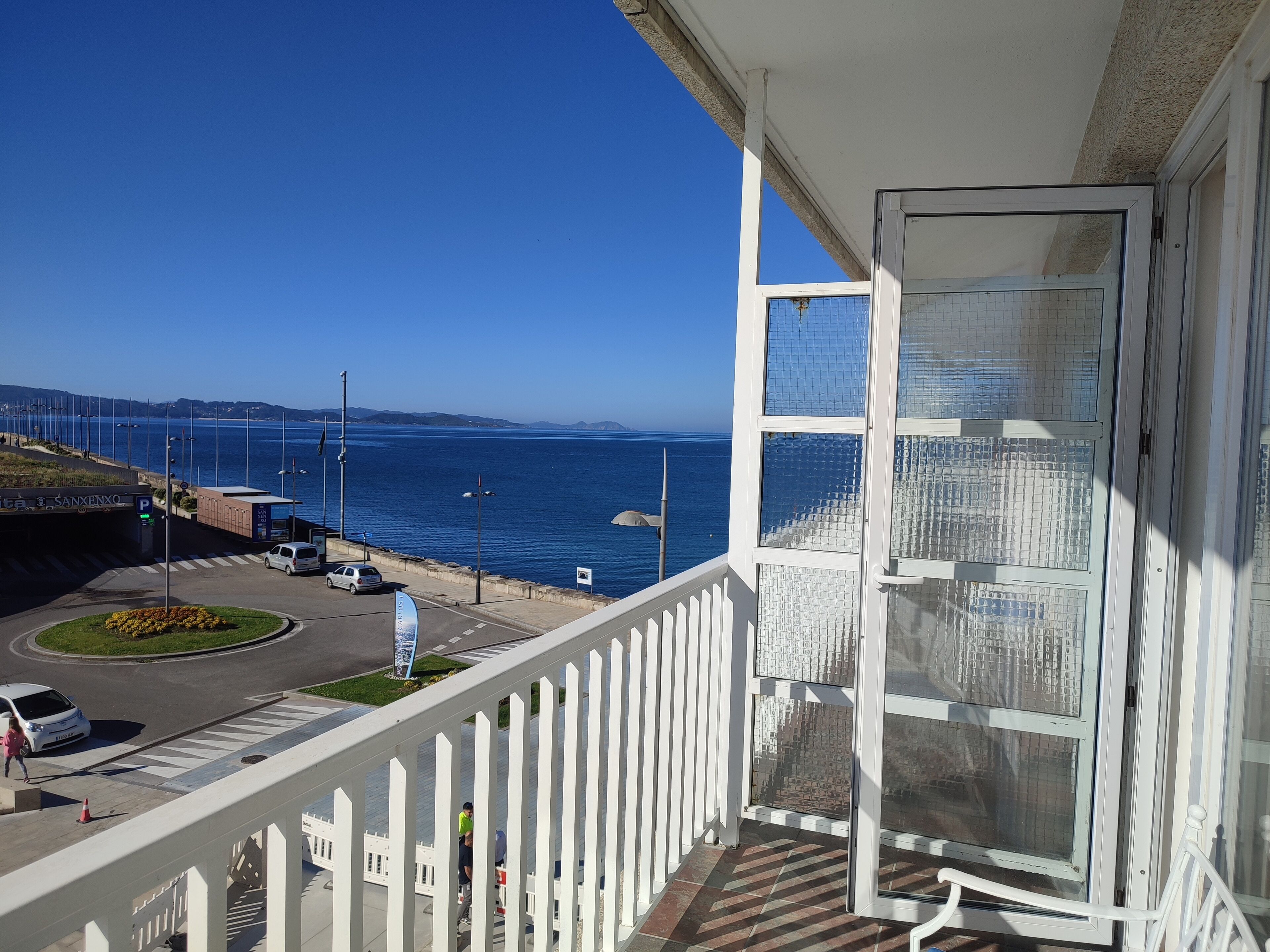 A balcony with a view of the sea, a parking area, and a building entrance.