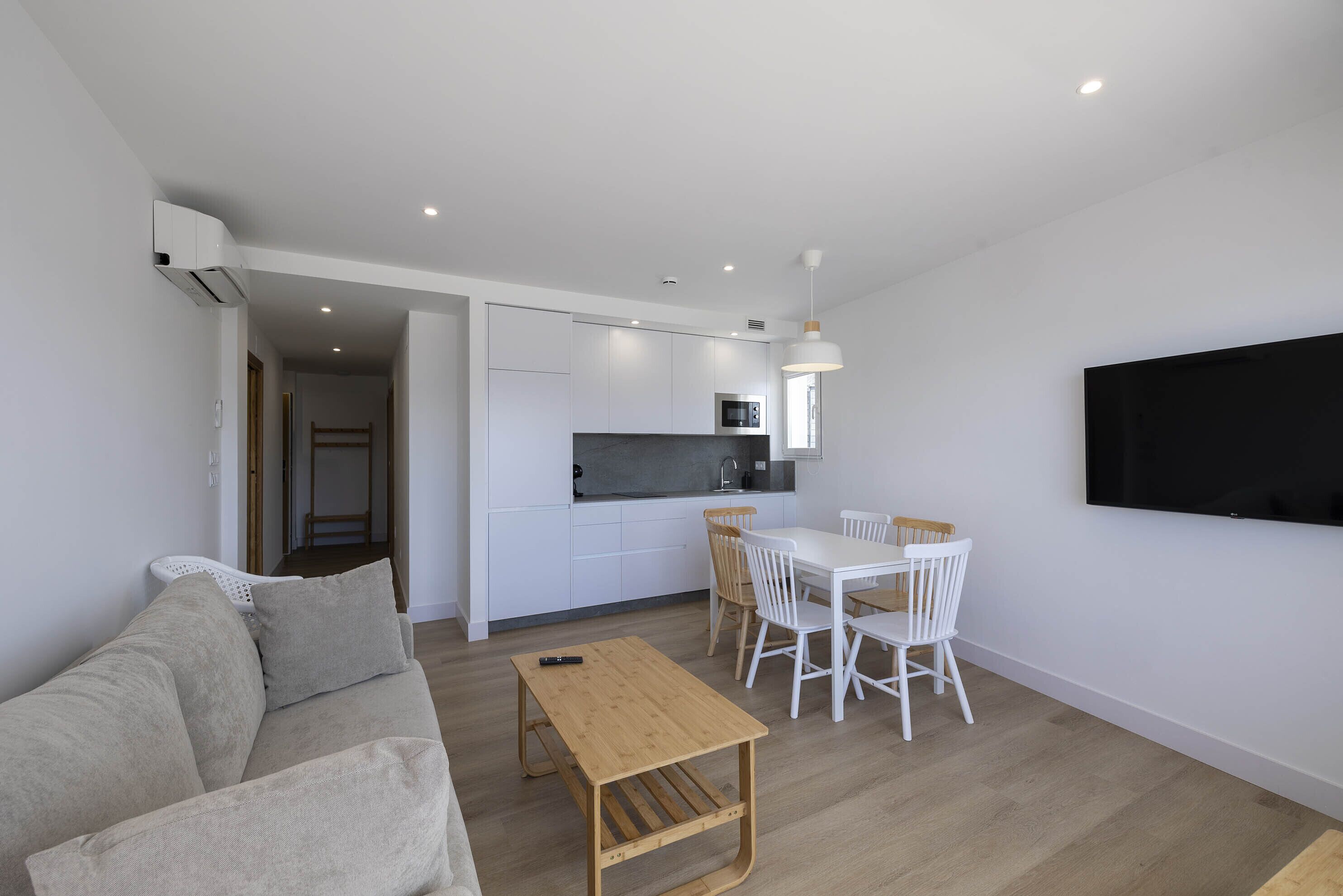 A modern living room with a sofa, a wooden coffee table, and a dining area with white chairs and a table. There is a kitchen area with white cabinets and a black countertop.