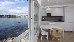 A modern kitchen with white cabinetry, a dining area with a white table and wooden chairs, and a large window overlooking a body of water with a small island and a boat. - Apartamentos Rotilio (Sanxenxo)
