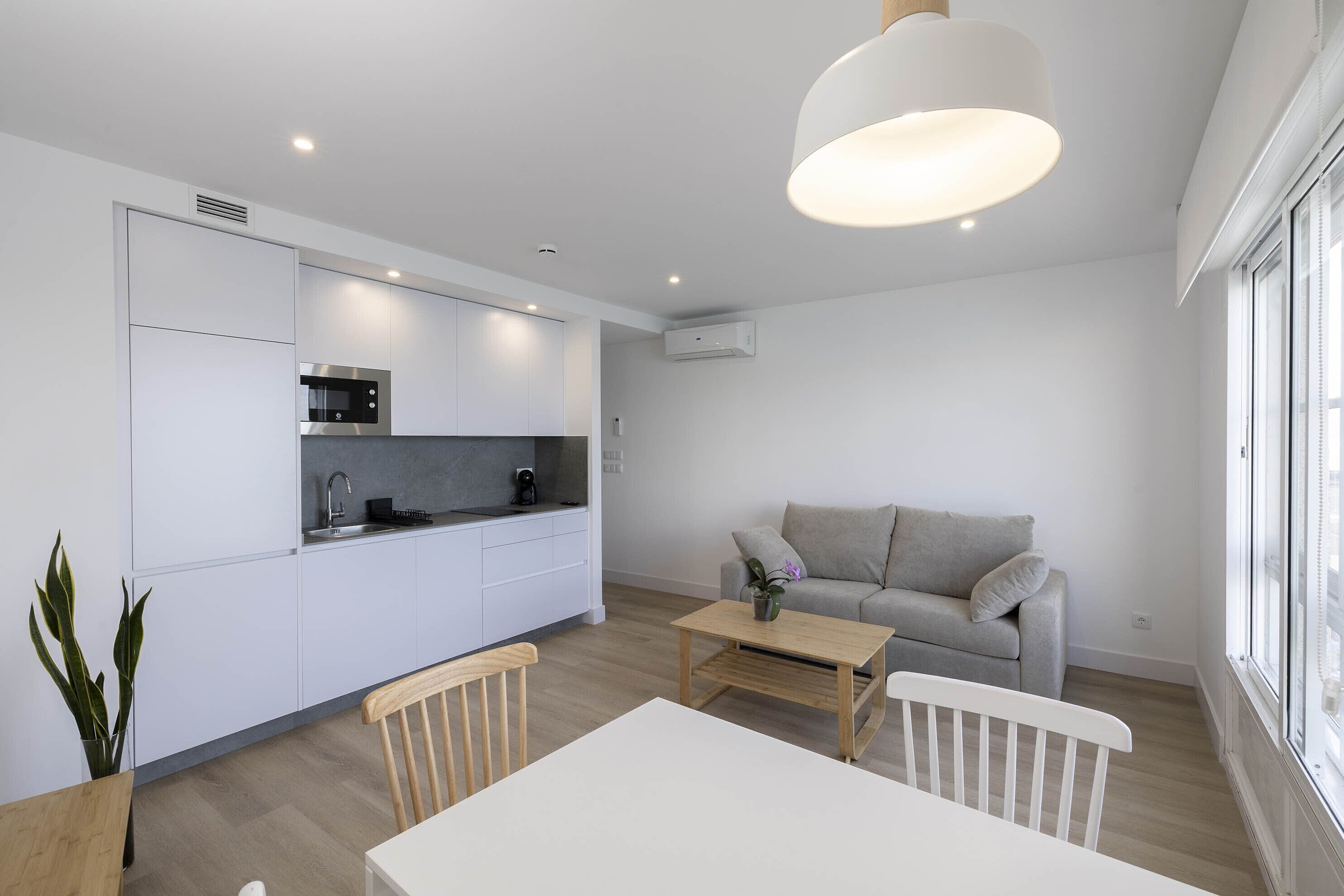 Una cocina moderna con armarios blancos, un comedor con mesa y sillas blancas, y un sofá gris en una sala de estar con un ventanal grande.