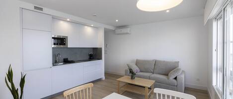 Una cocina moderna con armarios blancos, un comedor con mesa y sillas blancas, y un sofá gris en una sala de estar con un ventanal grande.