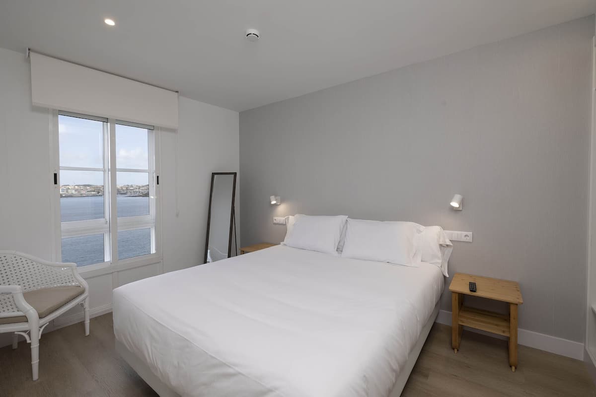 Quarto com cama grande, uma cadeira, uma mesinha e vista para o mar.