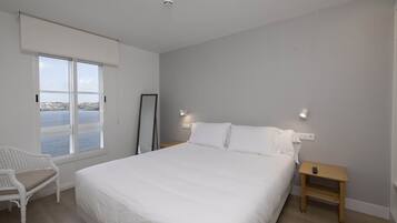 A bedroom with a large bed, a chair, a small table, and a view of the sea.