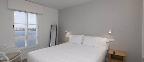 Quarto com cama grande, uma cadeira, uma mesinha e vista para o mar.