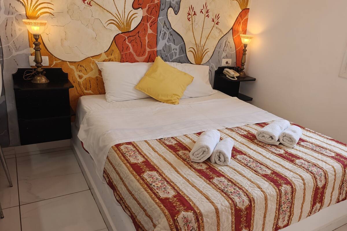 Un letto rifatto con una coperta a righe, due asciugamani arrotolati, un comodino con una lampada e una parete con un murale floreale.
