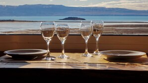 A table with four wine glasses and two plates, set against a scenic view of a beach and mountains. - Aikendor Hotel Panorámico (El Calafate)