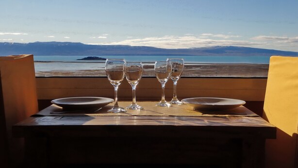 A table with four wine glasses and two plates, set against a scenic view of a beach and mountains. - Aikendor Hotel Panorámico (El Calafate)