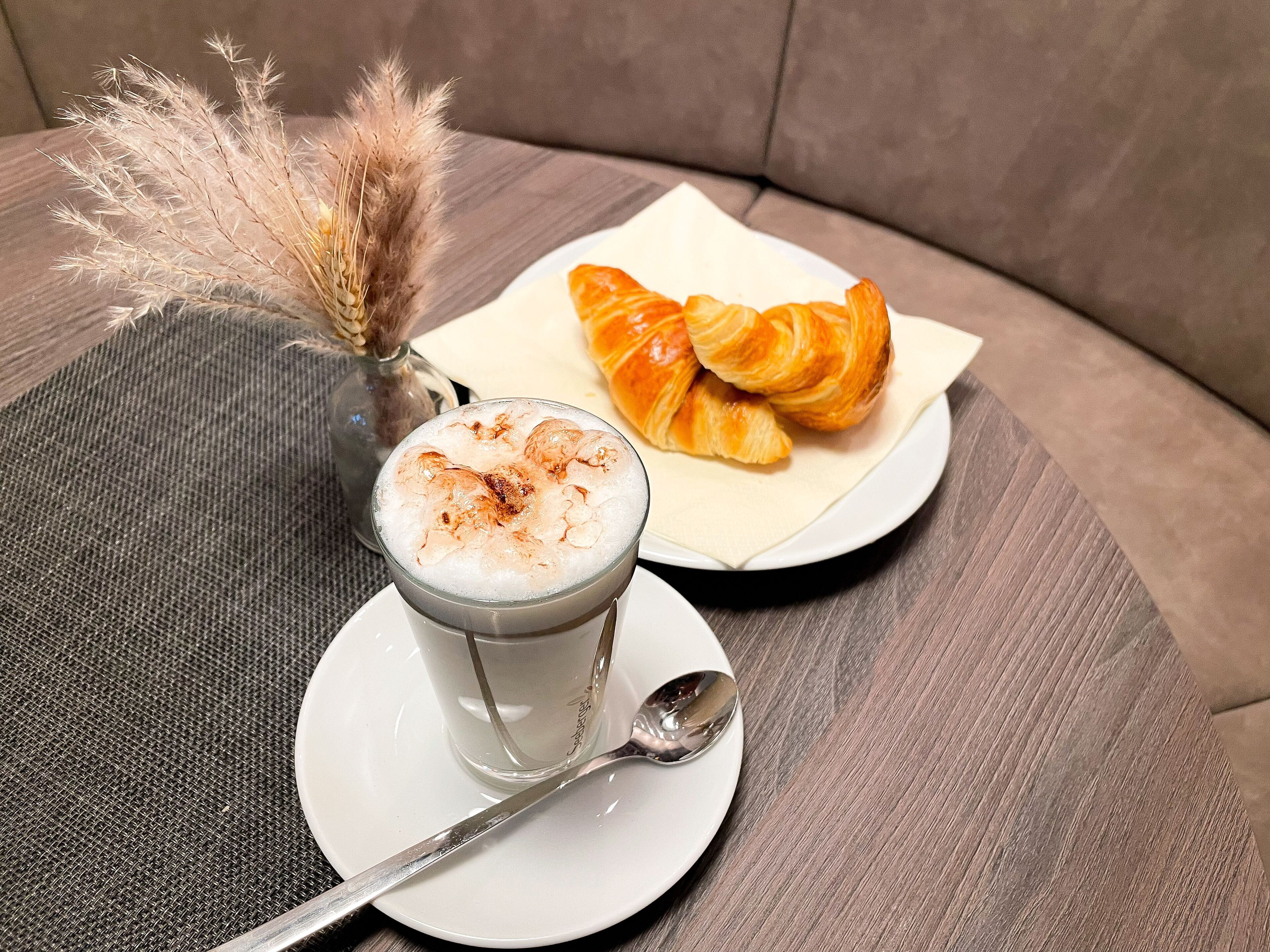 Ein Glas schaumiges Getränk mit einem Löffel auf einem weißen Teller, ein Croissant auf einem Serviettenstück und getrocknete Pflanzen auf einer strukturierten Oberfläche.