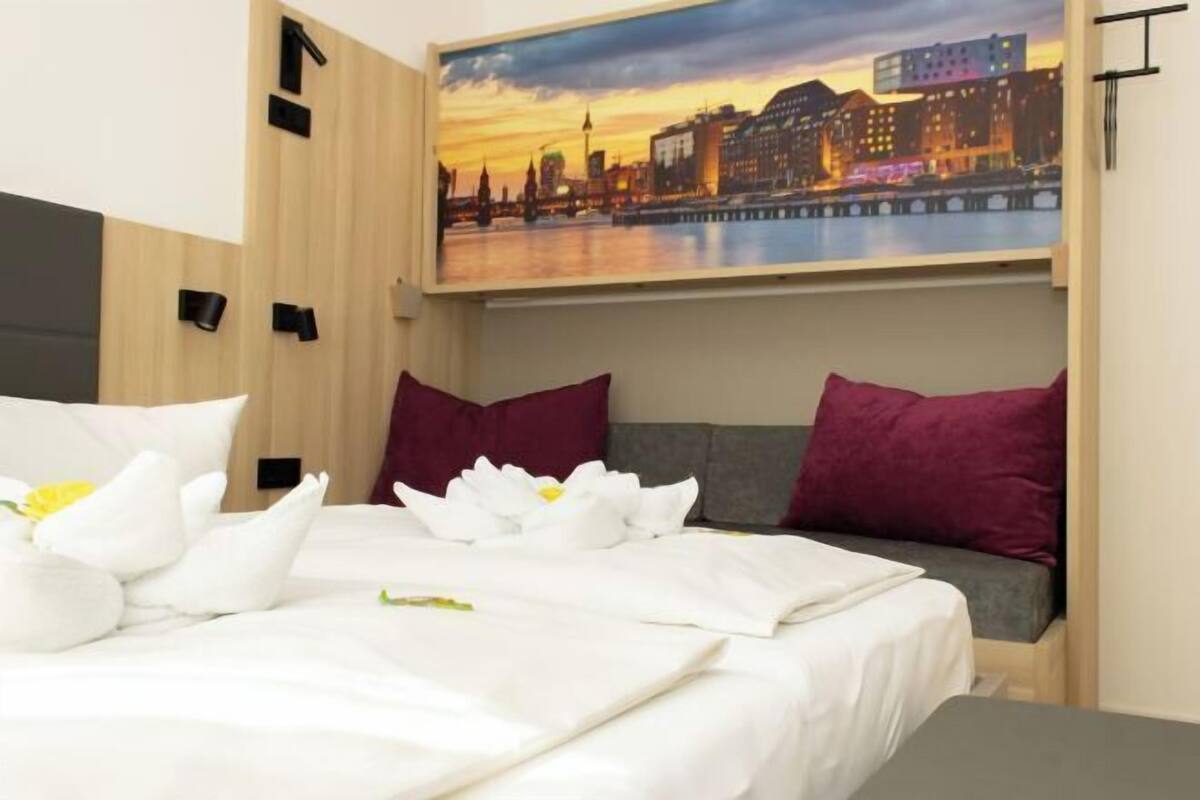 A neatly made bed with white linens and a floral arrangement, a cityscape painting above the bed, and a gray sofa with a purple pillow.