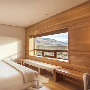 A bedroom with a large window, wooden walls, a bench, and a bed.