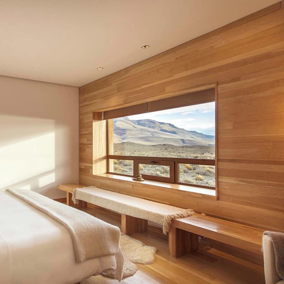 A bedroom with a large window, wooden walls, a bench, and a bed.