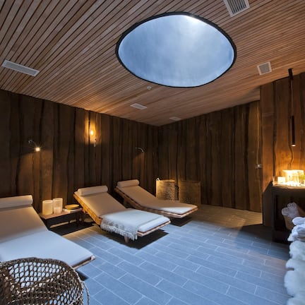 A spa room with wooden paneling, three sunbeds, a small table with candles, and a large circular skylight.