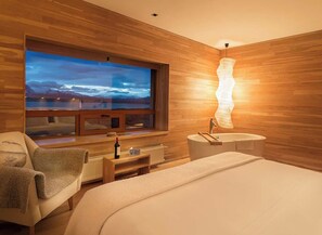 A modern bedroom with a large window, wooden walls, a bed, a chair, a nightstand, and a unique wall-mounted light fixture.