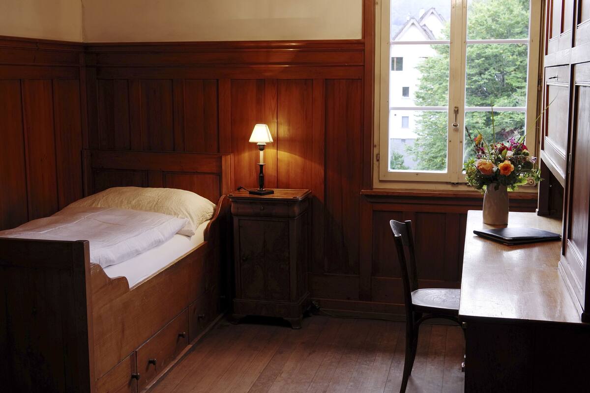 A bedroom with a wooden bed, a nightstand with a lamp, a chair, and a window with a view of buildings and trees.