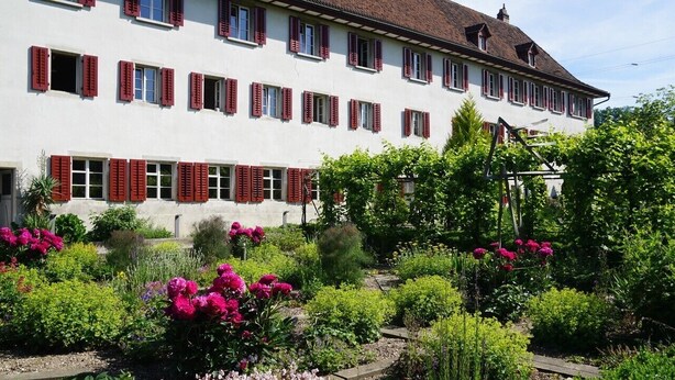 Ein großes, weißes Gebäude mit roten Fensterläden, umgeben von einem gepflegten Garten mit verschiedenen Pflanzen und Blumen.