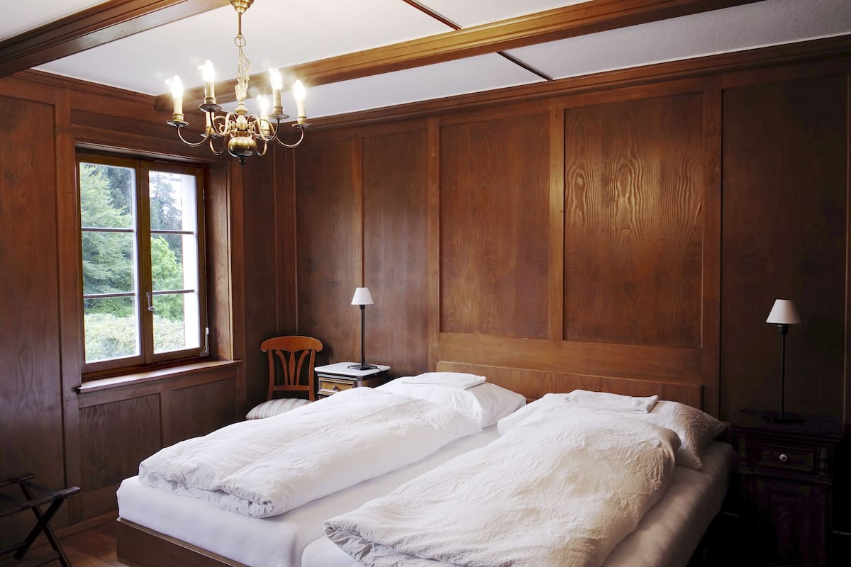 A room with two beds, wooden paneling, a chandelier, a window with a view of greenery, and a small table with a lamp.