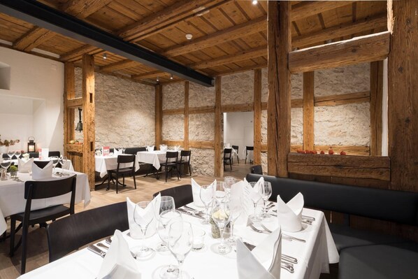 Un restaurante con vigas de madera y paredes de piedra, mesas dispuestas con manteles blancos y copas de vino.