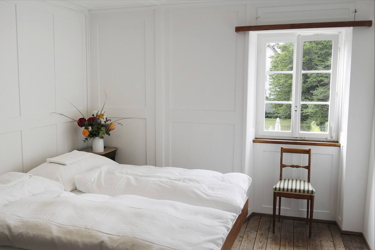 A bedroom with a bed, a chair, a window, and a vase with flowers.