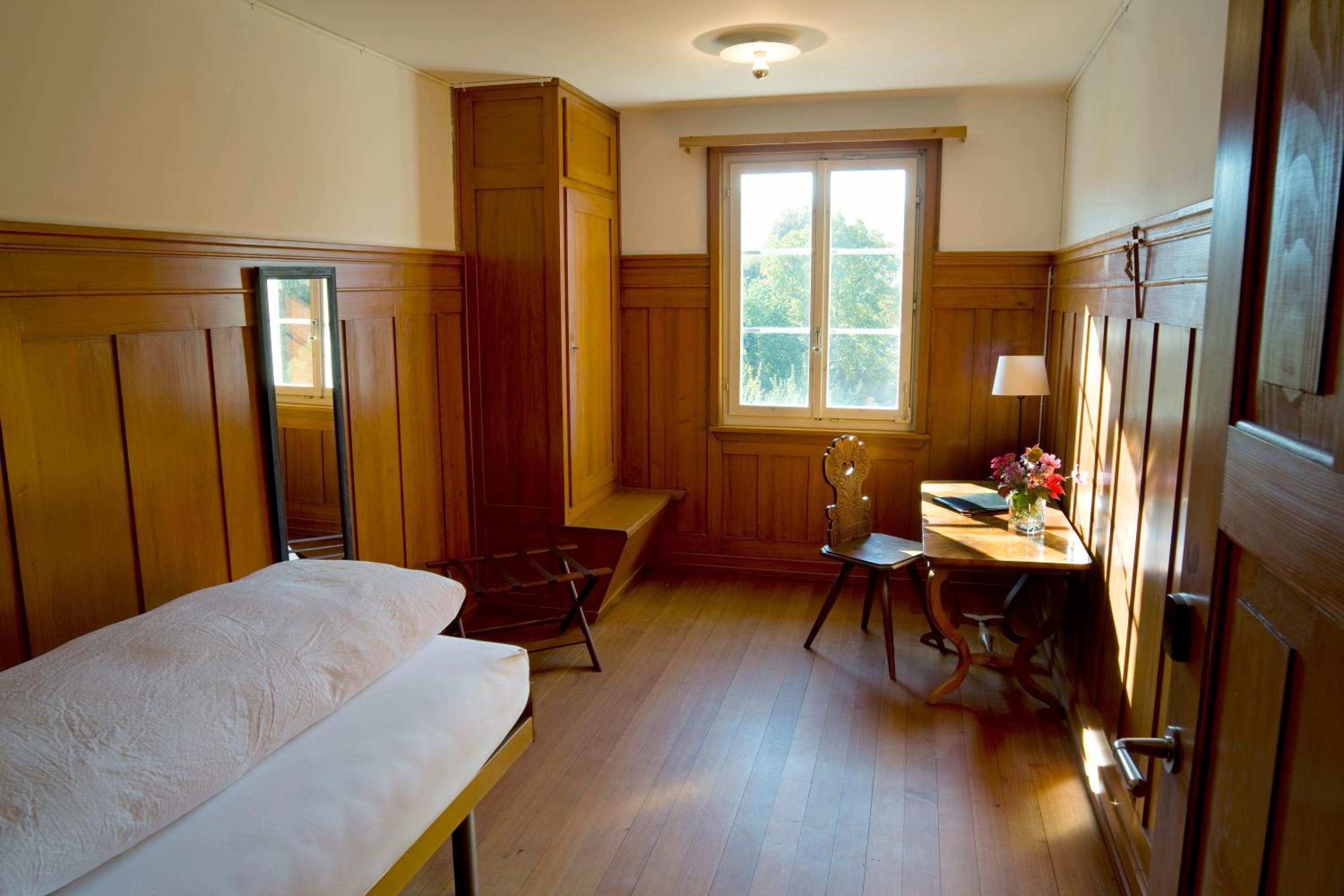 A room with wooden paneling, a bed, a desk with a lamp, and a window with a view of greenery.
