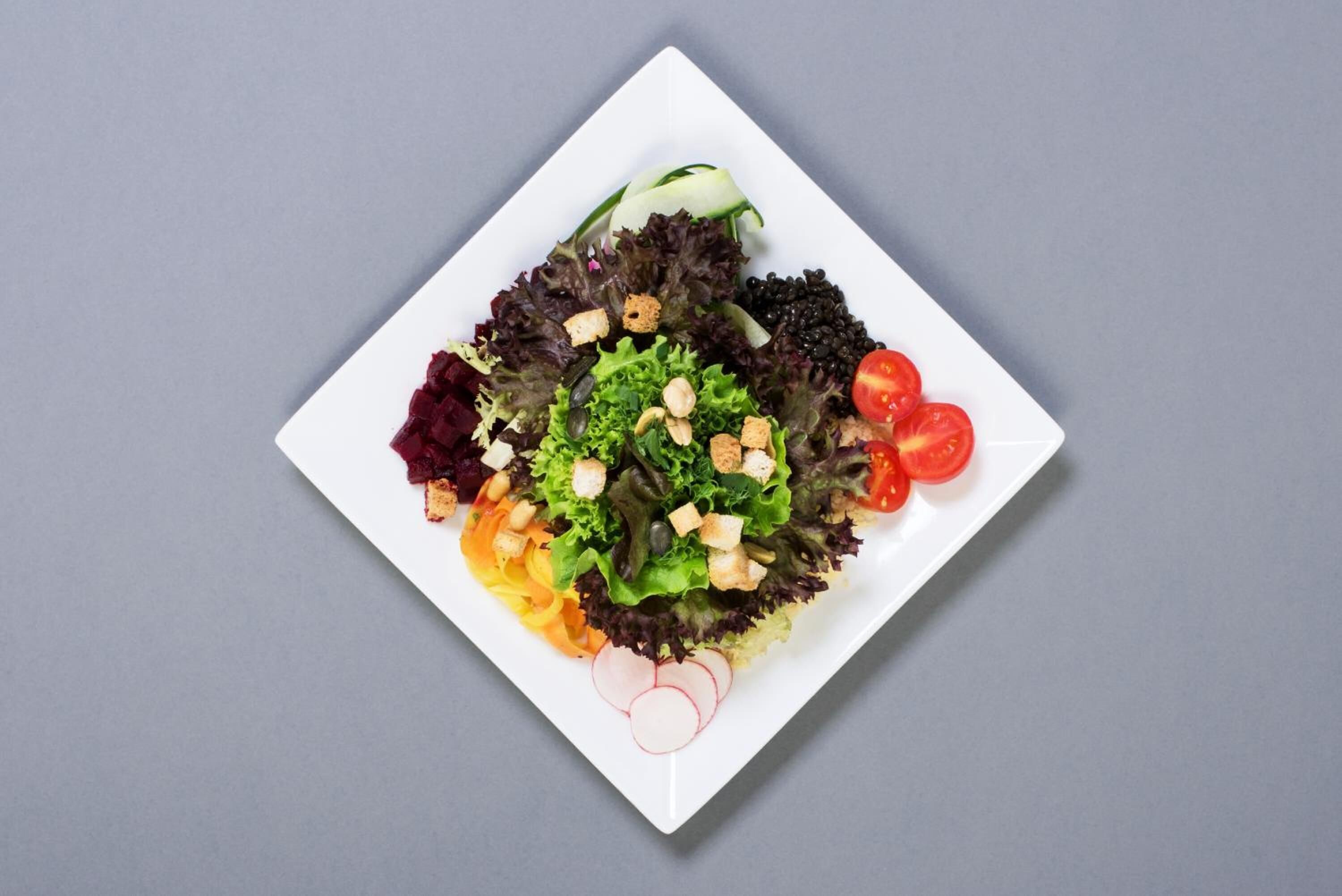 Ein weißes, quadratisches Teller mit einem Salat aus verschiedenen Gemüsesorten und Nüssen.