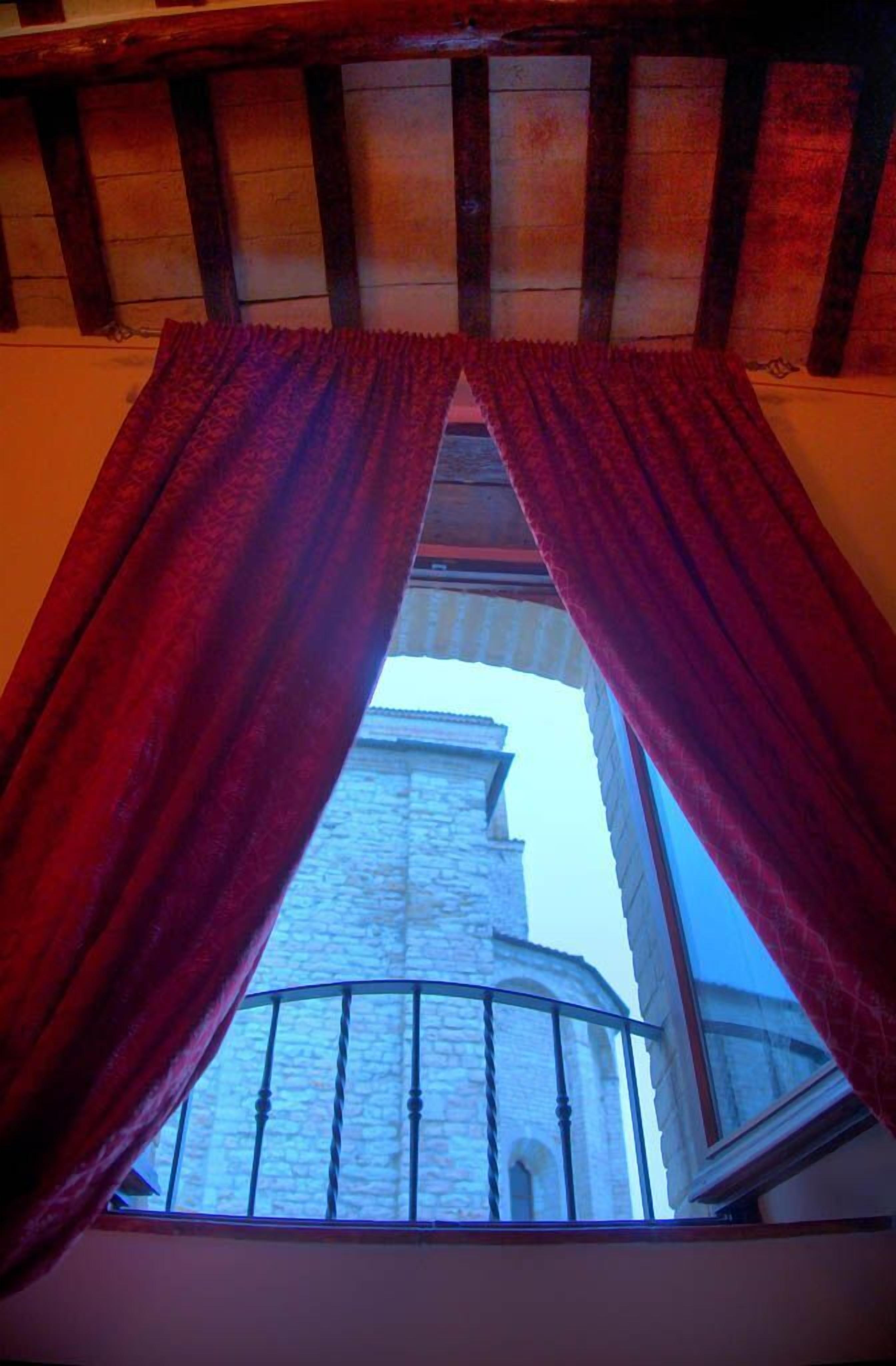 Vista attraverso una finestra con tende rosse, che mostra un edificio in pietra e un balcone con ringhiera in metallo.