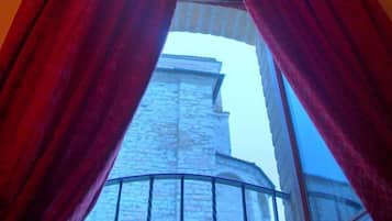 Vista attraverso una finestra con tende rosse, che mostra un edificio in pietra e un balcone con ringhiera in metallo.