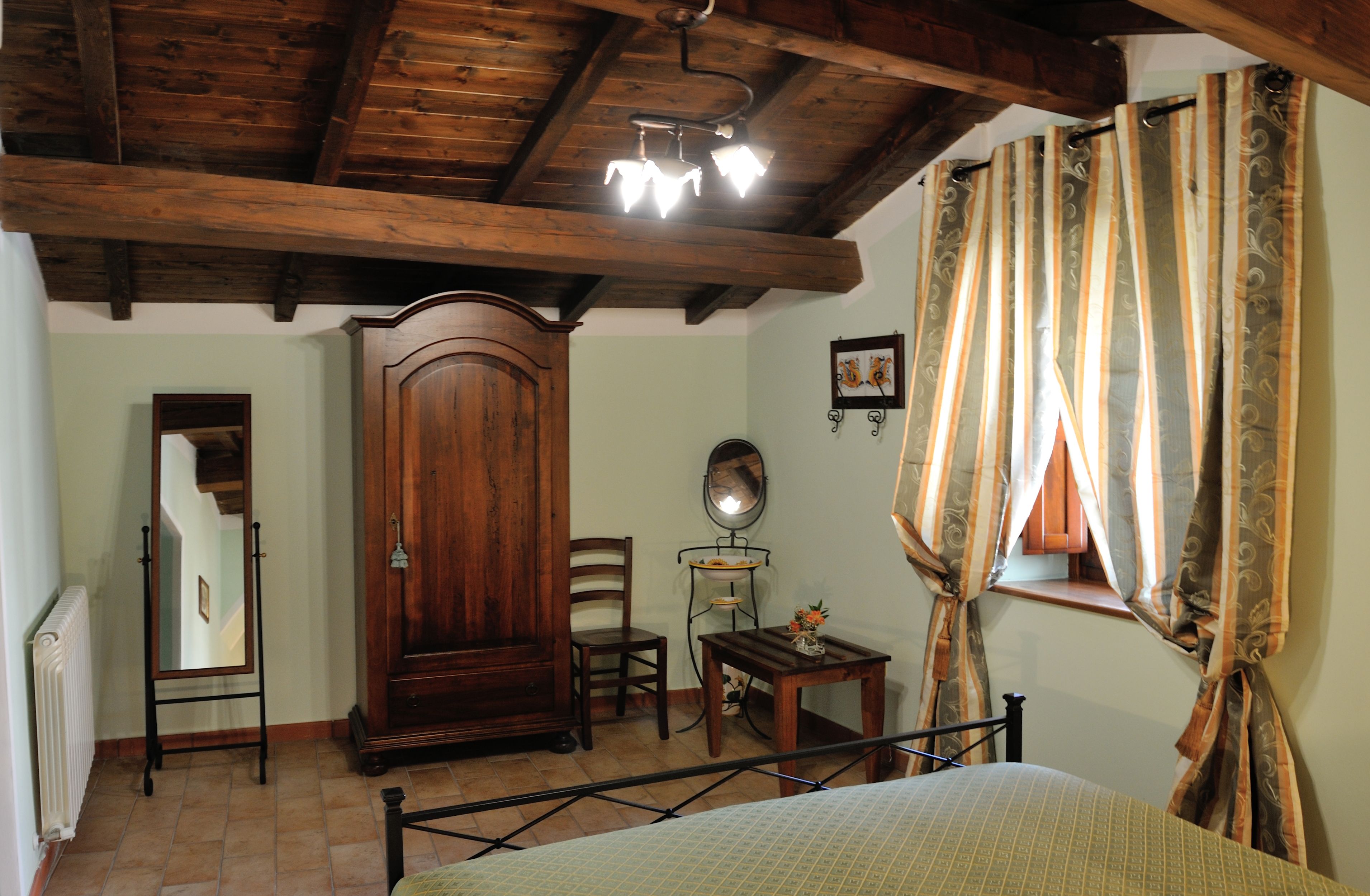 Ein Schlafzimmer mit Holzbalkendecke, einem Bett, einem Schrank aus Holz, einem Stuhl, einem kleinen Tischchen und Vorhängen.