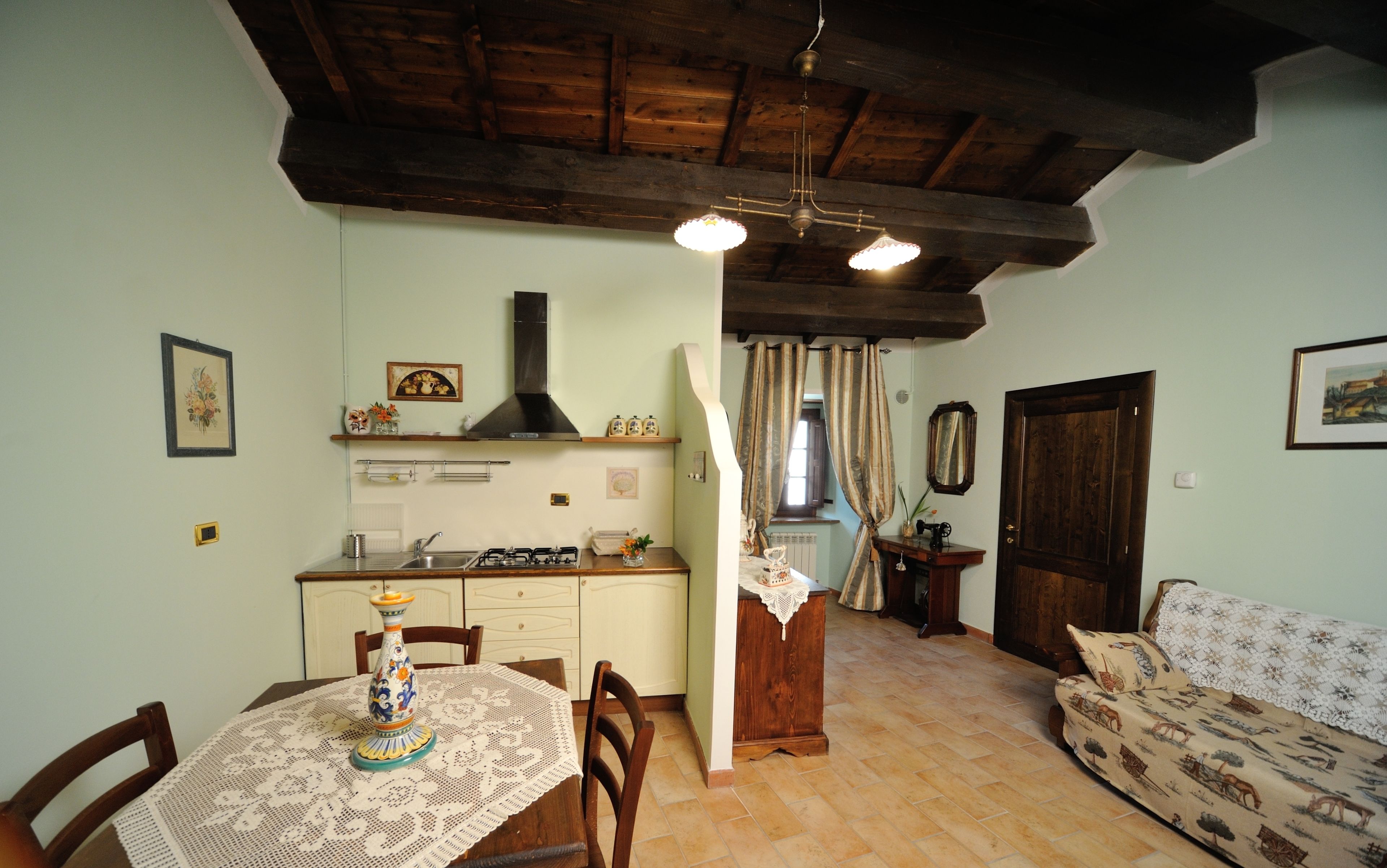Una sala de estar acogedora con un sofá, una mesa de comedor con mantel de encaje, y una zona de cocina con estufa a gas y armarios de madera.