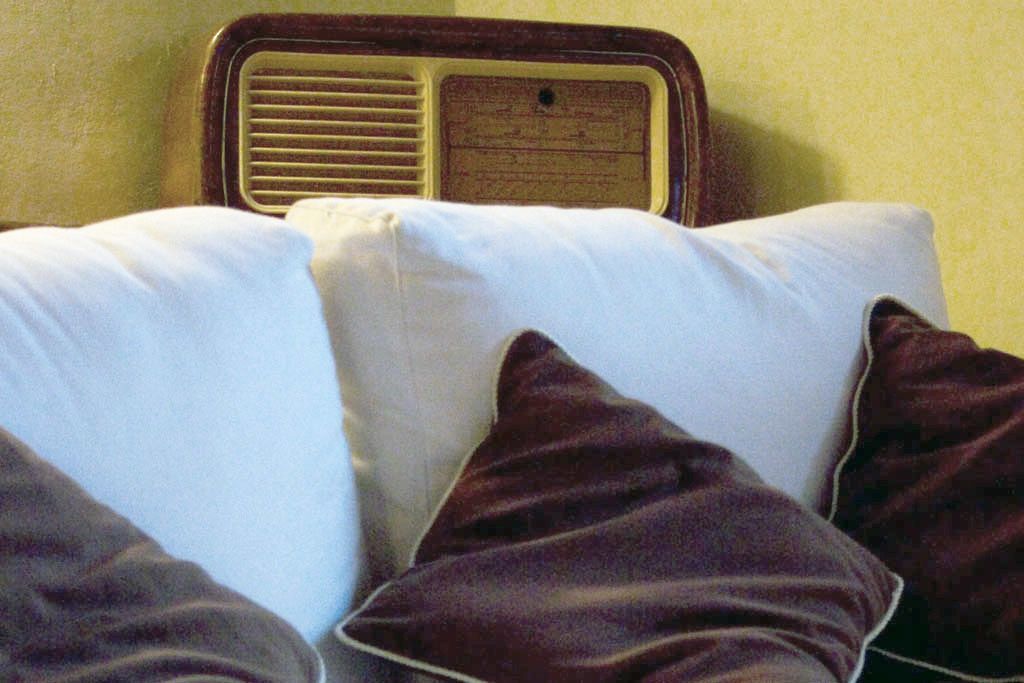 A bed with white and dark brown pillows, a wooden headboard with a radio, and a plain wall.