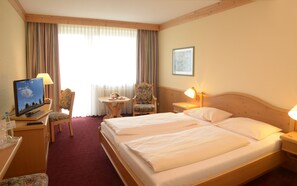 A hotel room with two beds, a desk with a computer, a chair, a small table with a lamp, and a window with curtains. - Hotel Kaiseralm (Bischofsgruen)