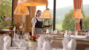 A restaurant with tables set for dining, a server attending to guests, and a scenic view through the window. - Hotel Kaiseralm (Bischofsgruen)