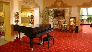 A grand piano in a room with red patterned carpet, a large mirror, and a chandelier. - Hotel Kaiseralm (Bischofsgruen)