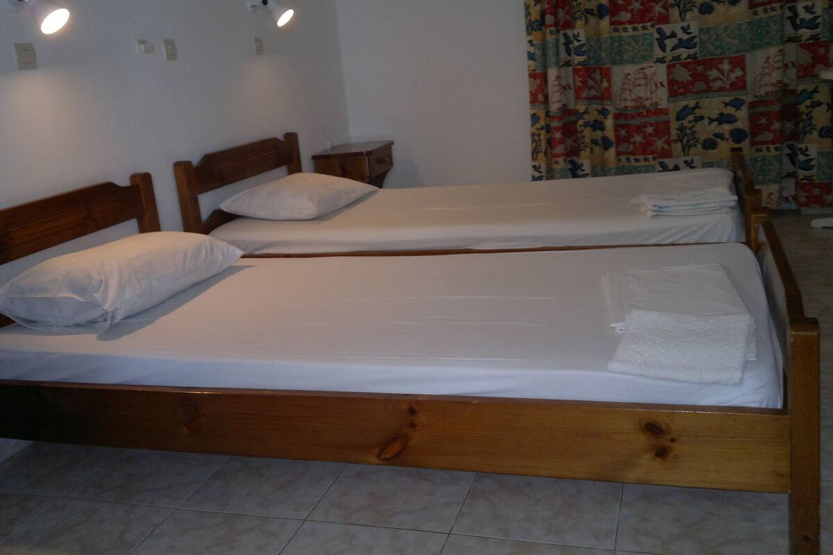 Two wooden beds with white linens, a small wooden nightstand, and a quilted curtain.