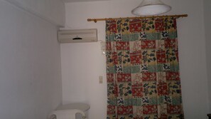 Un baño con un inodoro blanco, aire acondicionado instalado en la pared y una cortina de ducha de patchwork colorido.