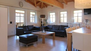 A spacious living room with a wooden ceiling, a black leather sofa, a wooden coffee table, and a dining table with chairs. - AvenidA Residenz Burg by Alpin Rentals (Kaprun)