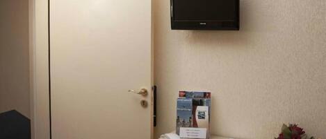 A hotel room with a TV mounted on the wall, a desk with a phone, a brochure, and a bouquet of flowers.