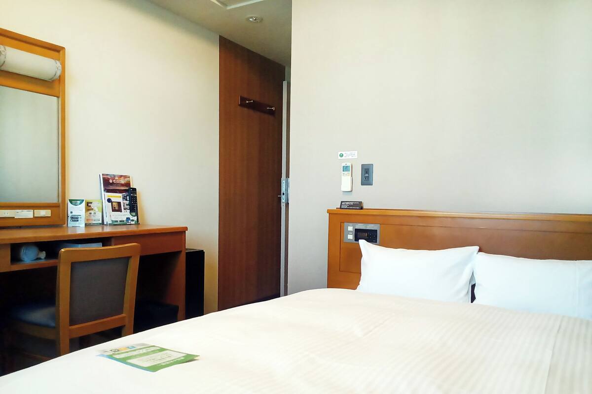 Camera d'albergo con un letto, una scrivania, una sedia, uno specchio, un comodino e un pannello di controllo a parete.