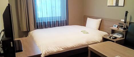 A hotel room with a bed, bedside table, and a desk. There is a window with curtains, a wall-mounted artwork, and a nightstand with a phone and lamp.