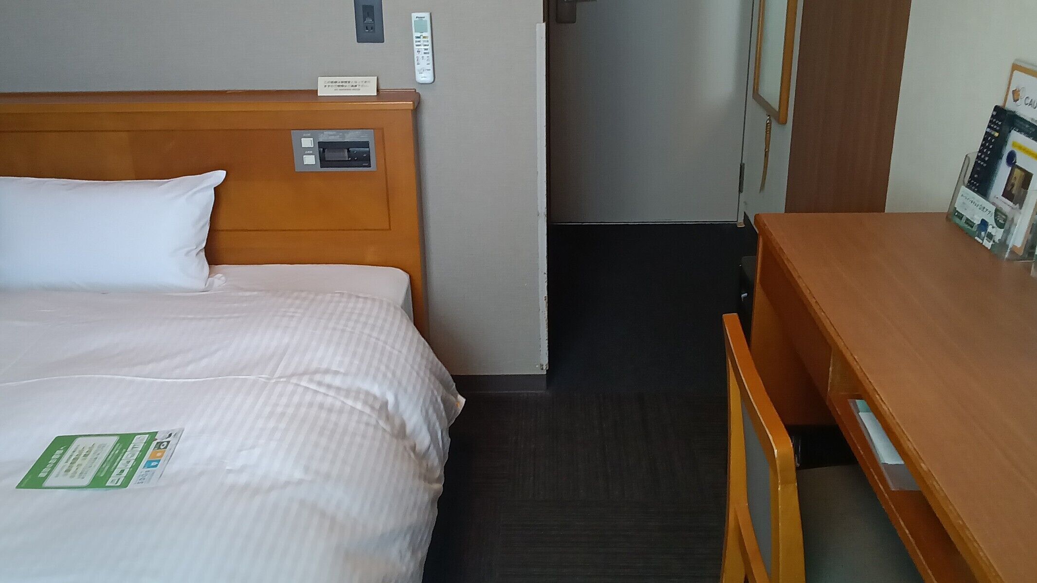 A hotel room with a bed, a wooden nightstand, a desk, a chair, and a door.